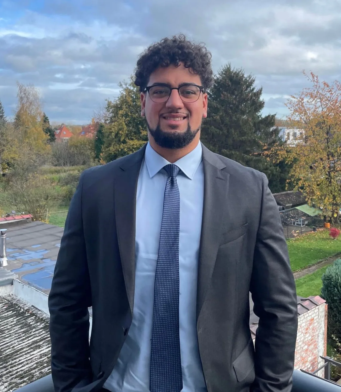 Sam Labenne, smiling, in a dark suit and tie on a balcony overlooking autumn trees